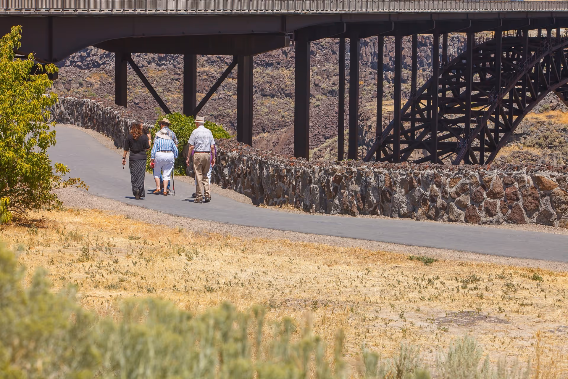 Four people walking on a paved path next to a stone wall with a large metal bridge structure in the background and dry grassland surrounding the path.
