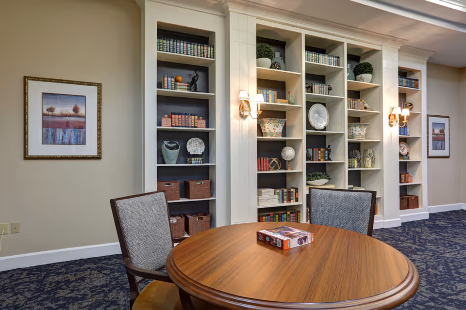 A cozy interior room with a round wooden table and two upholstered chairs. Behind the table is a wall with built-in shelves filled with books, decorative items, and baskets. The room has framed artwork on the walls and wall-mounted lamps providing warm lighting. The floor is carpeted with a patterned design.