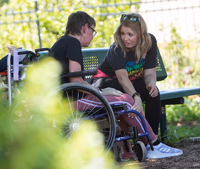 A woman sitting on a bench outdoors is attentively talking to a man in a wheelchair. They are surrounded by greenery and natural light.