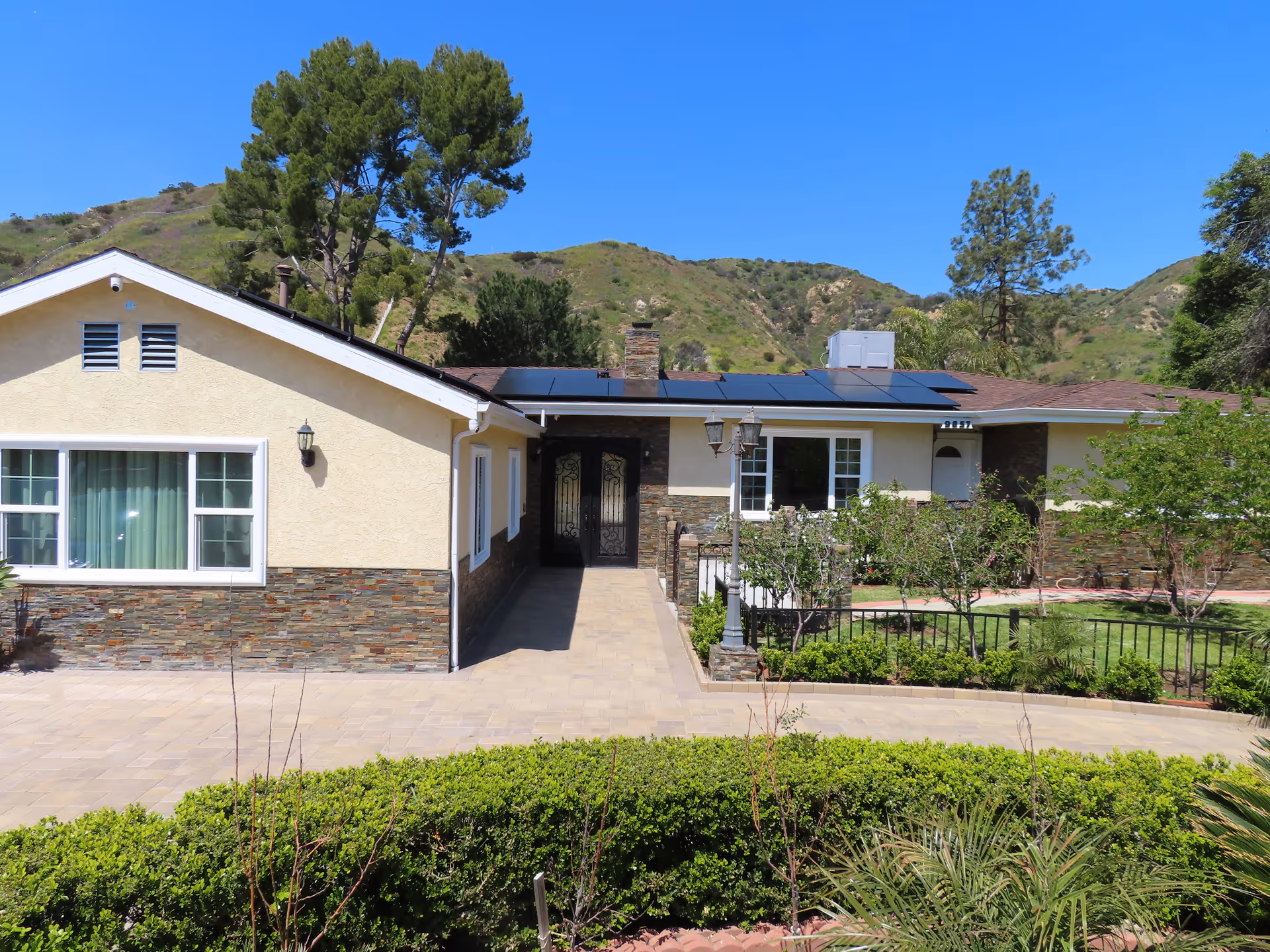 Single-story home front with a paved driveway, landscaped hedges, and solar panels on the roof against a hillside backdrop.