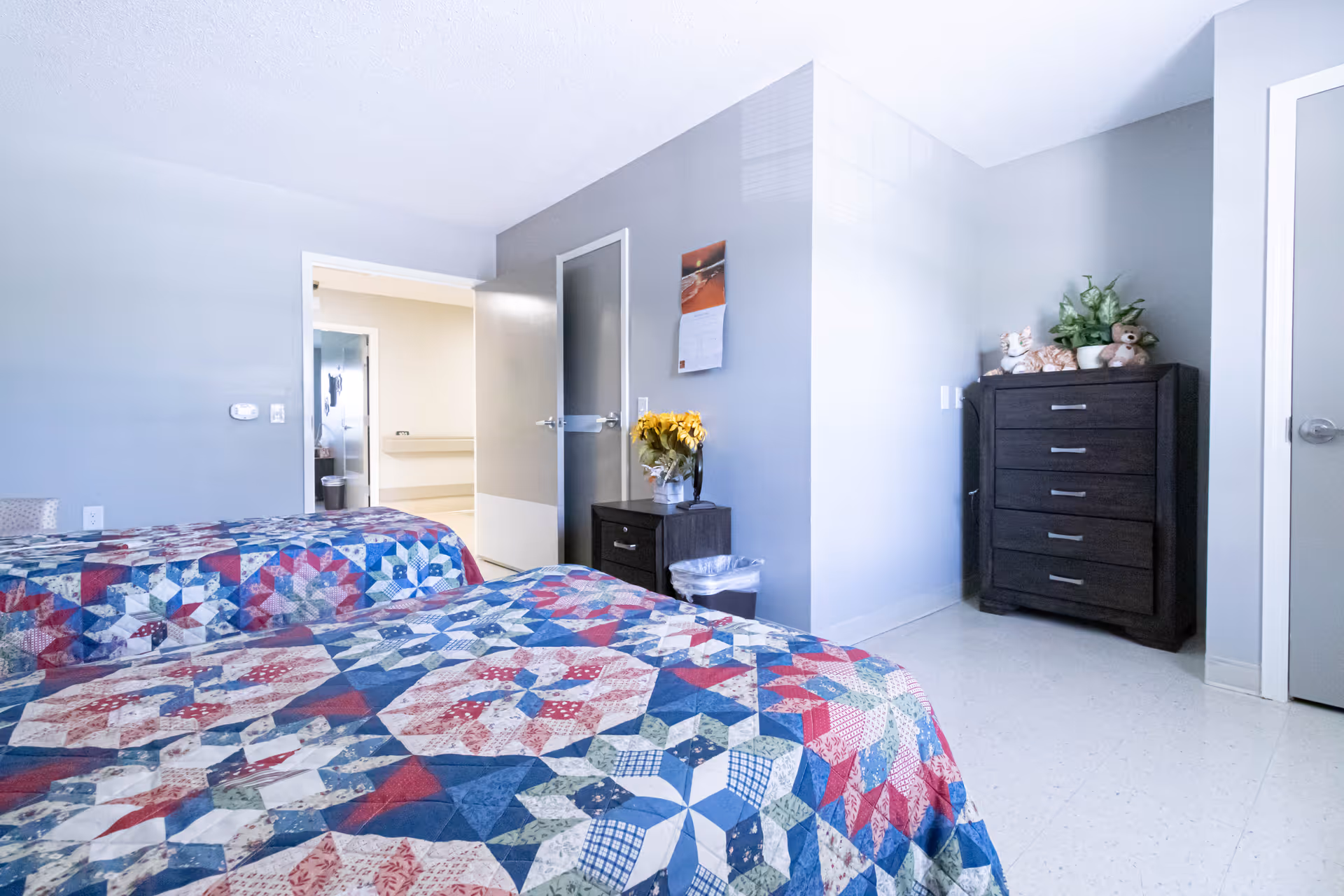 A bright bedroom with two twin beds covered in colorful patchwork quilts. The room has light gray walls, a small dark wooden nightstand with a vase of yellow flowers, and a dark wooden dresser with stuffed animals and a plant on top. There are two doors, one open leading to a hallway and another closed. The floor is light-colored and the room is well-lit with natural light.