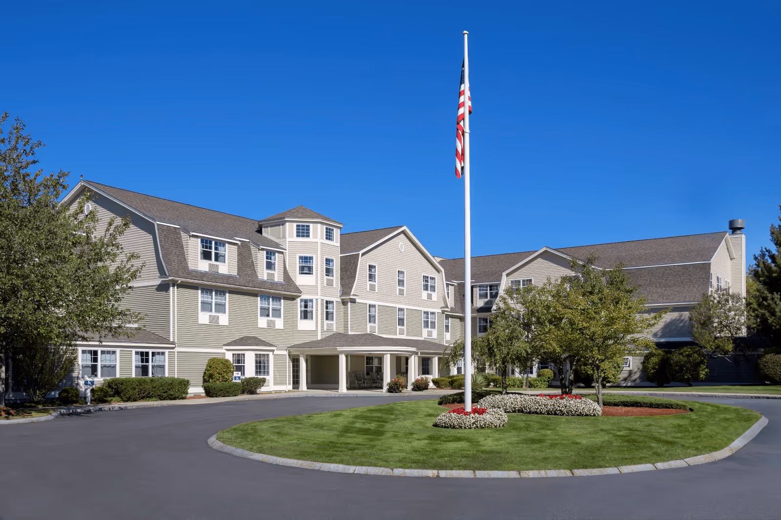 Exterior view of a large senior living facility building with beige siding and multiple windows under a clear blue sky. In front of the building is a circular driveway with a grassy island in the center, featuring a flagpole with an American flag and landscaped flower beds. Trees and shrubs surround the area.
