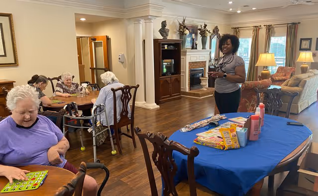 A senior living facility common area with elderly residents sitting at tables playing bingo and a staff member standing nearby. The room has wooden floors, a fireplace, a TV, and comfortable seating with large windows letting in natural light.