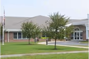 Single-story brick building with a covered entrance and circular drive, a grassy lawn with two small trees and an American flag nearby.