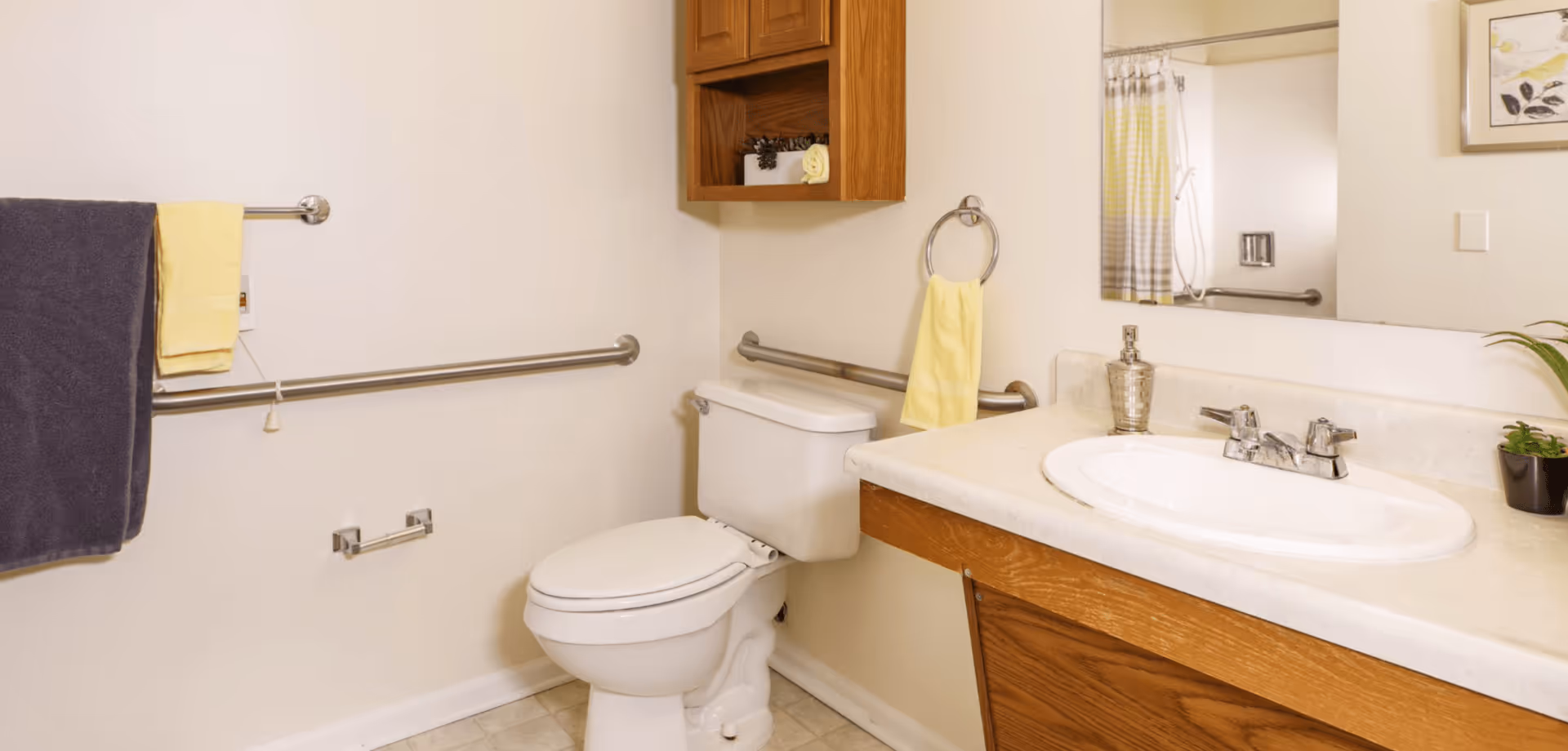A clean bathroom with a white toilet, a sink with a countertop, a large mirror above the sink, and a wooden cabinet mounted on the wall. There are grab bars installed near the toilet and a towel rack with a dark blue and a yellow towel. A yellow hand towel hangs on a ring next to the sink. The floor is tiled and the walls are light-colored.