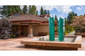 Outdoor courtyard area with a water feature consisting of two tall green glass sculptures in a rectangular pool. The area is paved with stone tiles and surrounded by trees and shrubs, with a building entrance visible in the background under a partly cloudy sky.