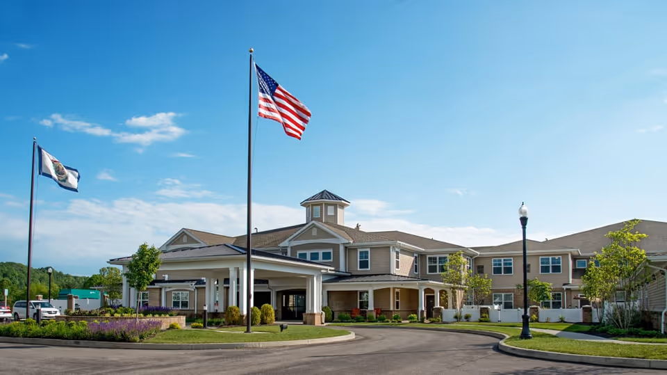 Exterior view of Bellaire at Devonshire Assisted Living & Memory Care building with two flagpoles displaying the American flag and another flag, surrounded by landscaped greenery and a clear blue sky.