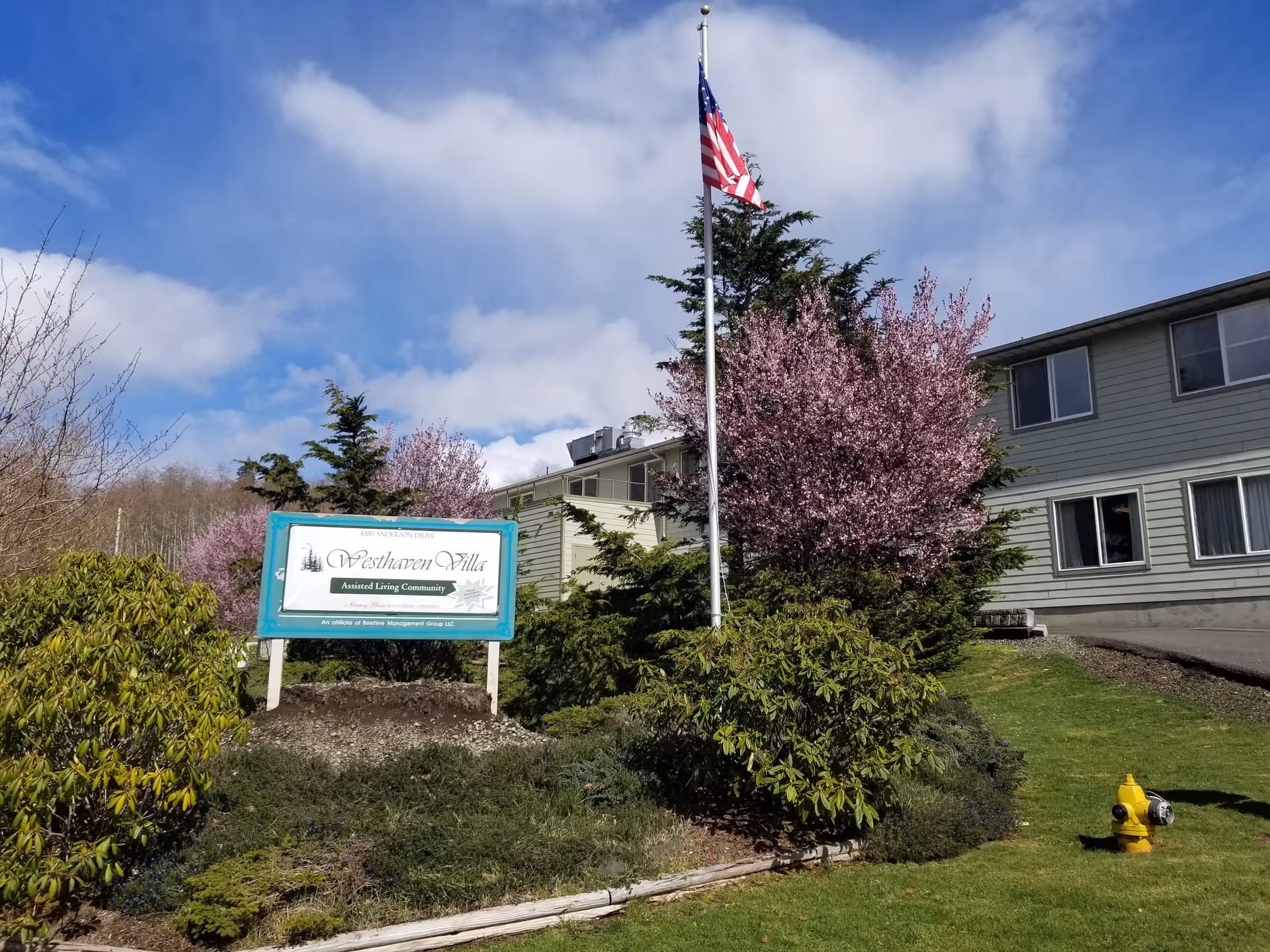 Exterior view of Westhaven Villa Assisted Living facility with a green and white sign in front, an American flag on a flagpole, blooming pink trees, bushes, and a yellow fire hydrant on a grassy lawn under a partly cloudy blue sky.