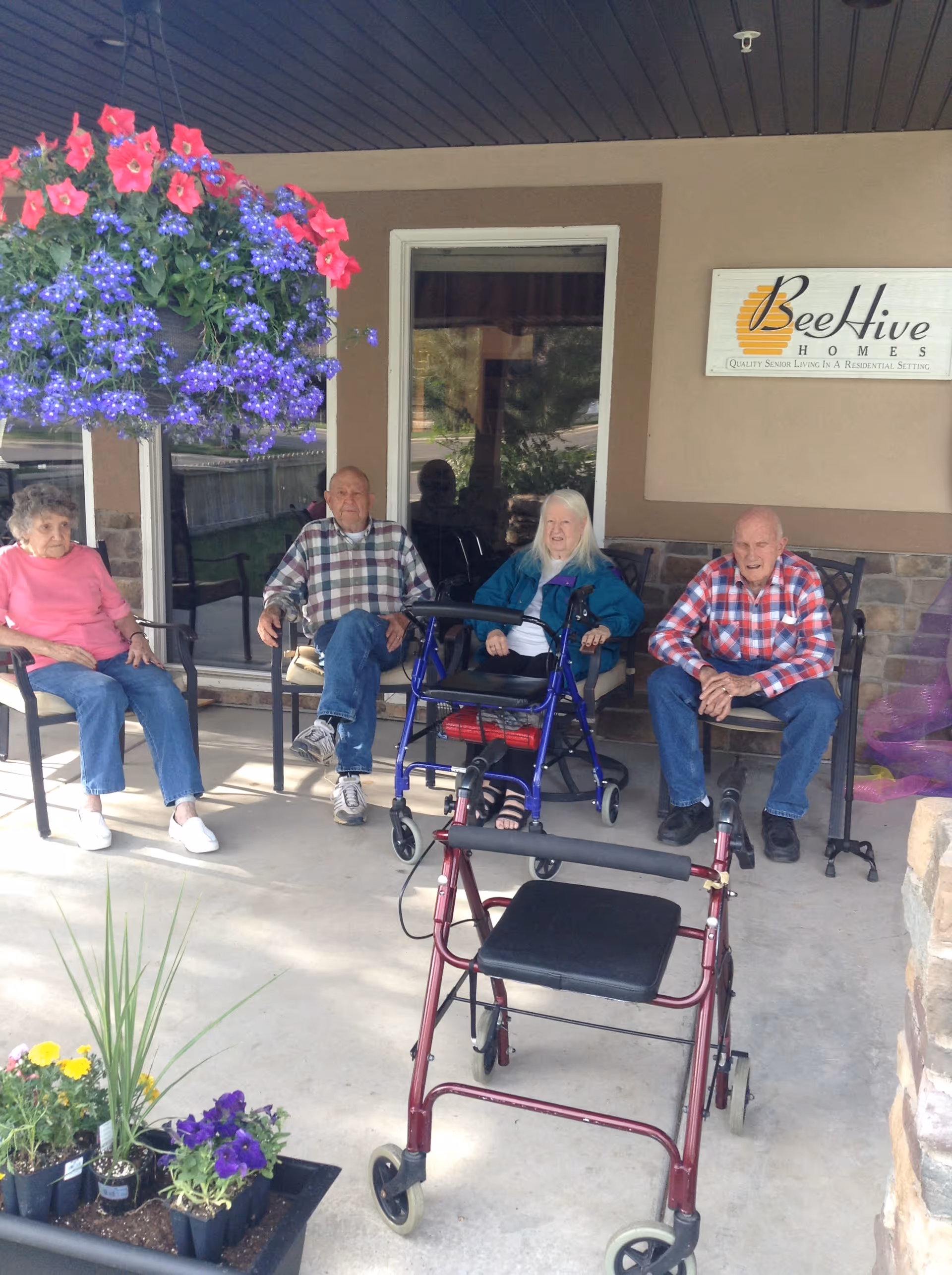 Four elderly people sitting on chairs outside a building under a covered porch. There are two walkers in front of them and hanging flower baskets with purple and red flowers. A sign on the wall reads 'BeeHive Homes Quality Senior Living In A Residential Setting.'