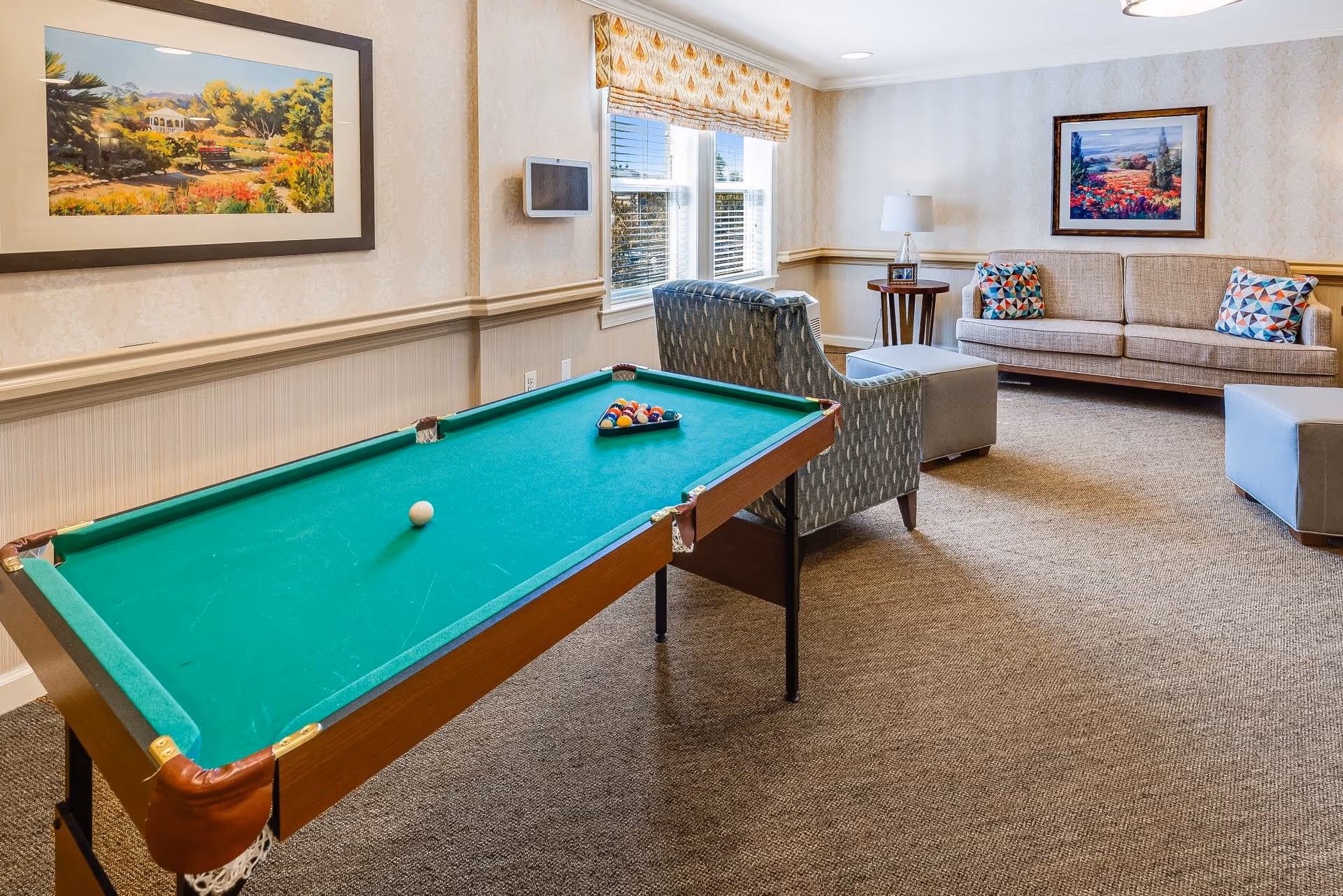 A cozy common room with a pool table in the foreground, a patterned armchair, a beige sofa with colorful geometric pillows, two ottomans, a side table with a lamp, and framed landscape paintings on the walls. The room has carpeted flooring and a window with a patterned valance.