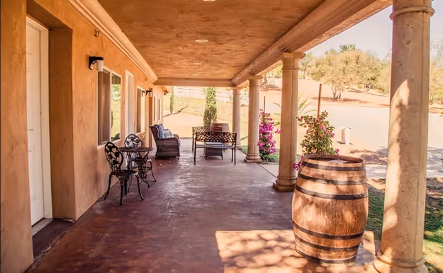 Covered porch with columns, metal bistro set, bench seating, potted plants, and a wooden barrel.