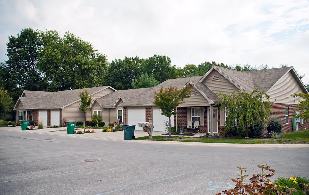 Row of single-story brick and siding assisted living units with garages, driveways, trash bins, and landscaping under a cloudy sky.