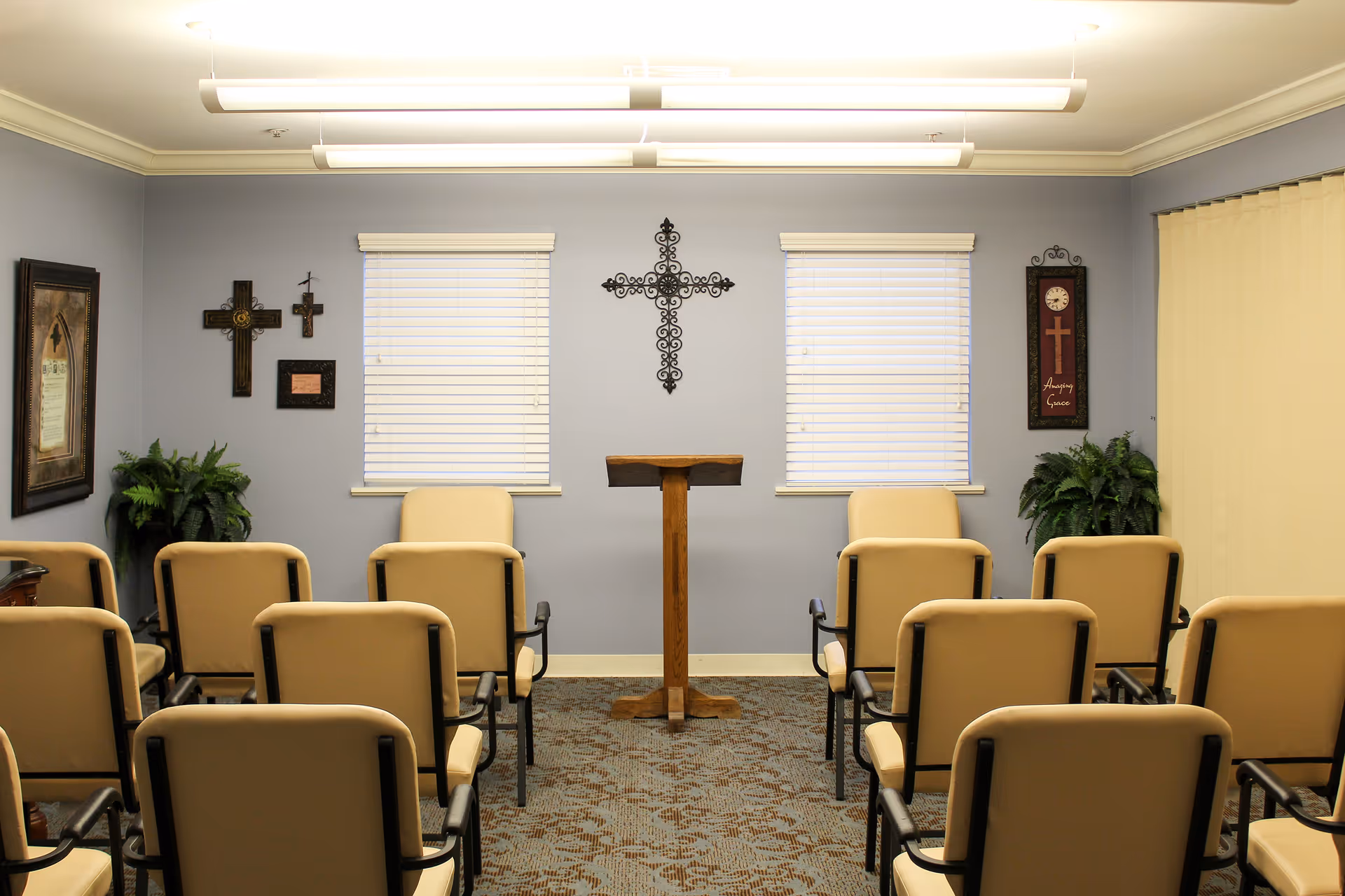 A small chapel or prayer room with beige chairs arranged in rows facing a wooden podium. The walls are light blue with two windows covered by white blinds. Several decorative crosses and framed religious artwork hang on the walls. There are two green plants in the corners of the room and overhead fluorescent lighting.