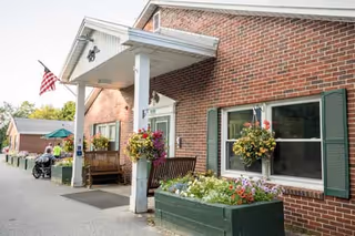 Exterior view of a brick residential care facility with a covered entrance supported by white columns. There are hanging flower baskets and a large planter with colorful flowers near the entrance. An American flag is mounted on the building. A person in a wheelchair is visible near the entrance.
