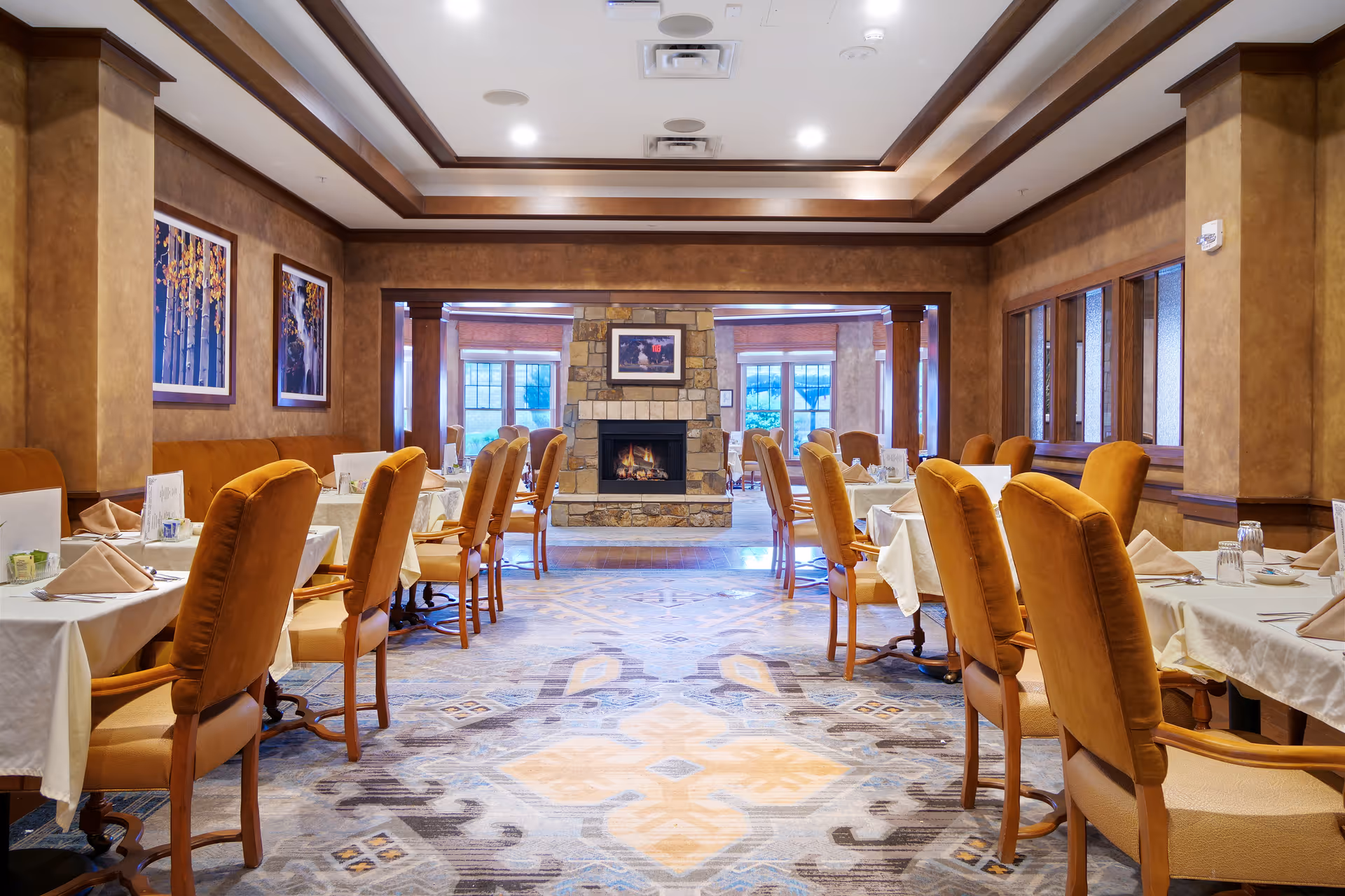 A warm, spacious dining room with neatly set tables and upholstered chairs facing a stone fireplace at the far end.