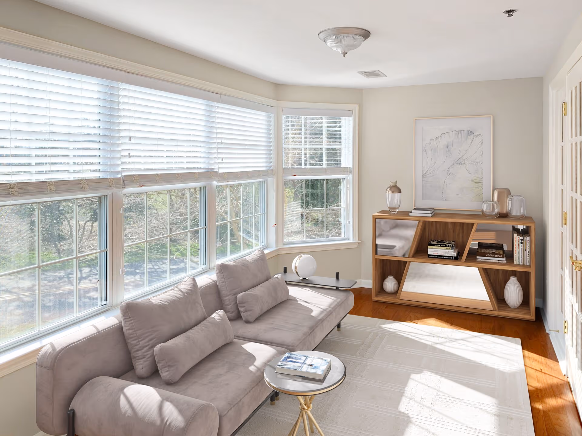A bright living room with large windows covered by white blinds letting in natural light. The room features a light gray sofa with matching cushions, a small round side table with books on it, a wooden bookshelf with decorative items and books, and a framed artwork on the wall. The floor is wooden with a light-colored area rug.