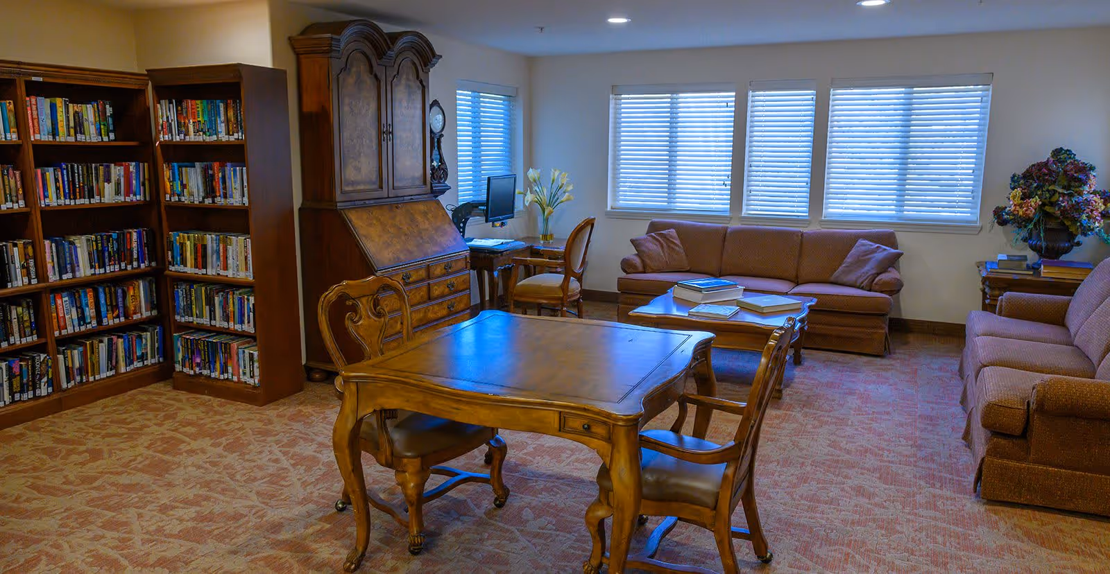 A cozy common room with bookshelves, a wooden table and chairs, sofas, and a computer desk by windows.