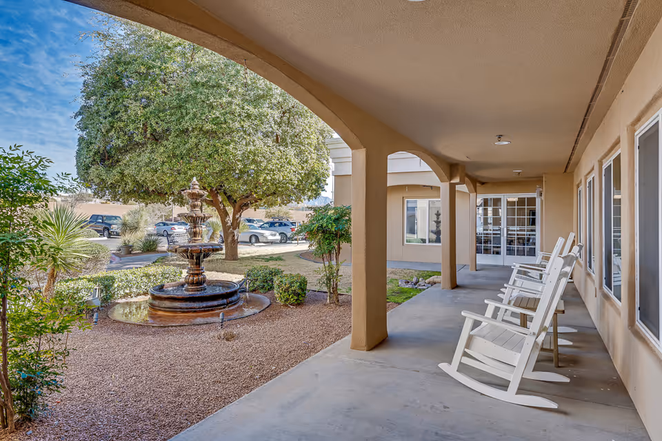 Covered outdoor patio area with several white rocking chairs lined up along the wall, overlooking a landscaped garden with a multi-tiered water fountain, trees, and shrubs. Several parked cars are visible in the background under a blue sky.