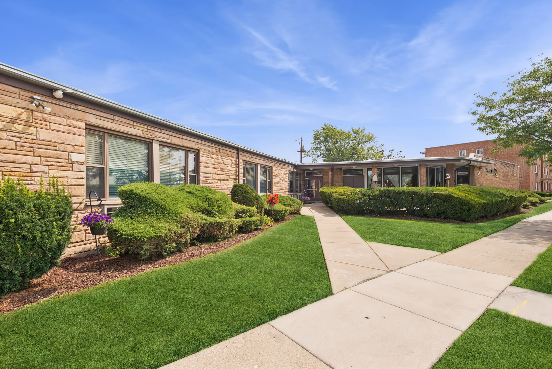 One-story brick building front with a central sidewalk, trimmed hedges and a green lawn under a blue sky.