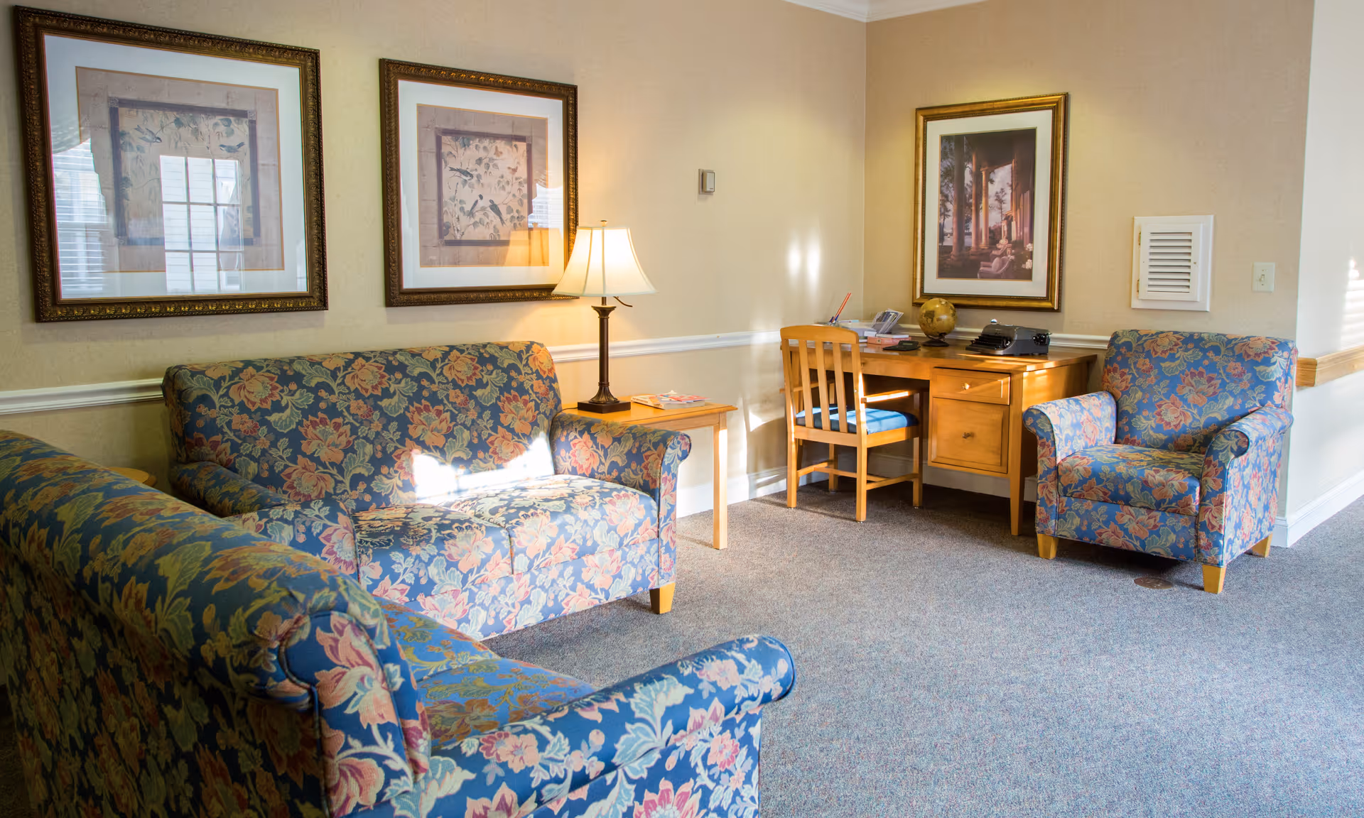 A cozy sitting area with floral patterned upholstered furniture including a sofa, loveseat, and armchair. There is a wooden desk with a chair, a lamp on a side table, and framed artwork on the walls. The room has beige walls and carpeted flooring.