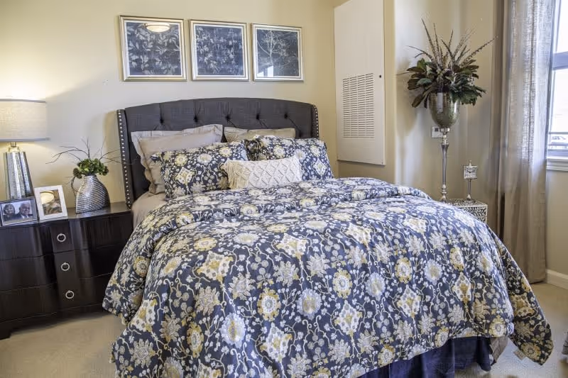 A neatly made bed with a dark upholstered headboard and floral patterned bedding in shades of blue, white, and yellow. On the left side, there is a dark wooden nightstand with a lamp, a small plant, and framed photos. Above the bed, three framed botanical prints hang on the wall. On the right side, a tall decorative vase with flowers stands near a window with beige curtains.