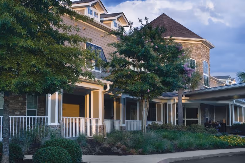 Exterior view of a senior living facility building with a covered porch, white railings, and landscaped greenery including trees and bushes under a partly cloudy sky.