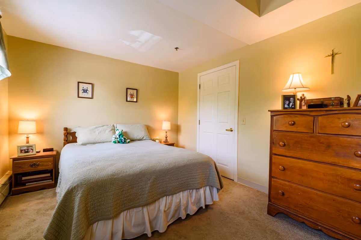 Well-lit bedroom with a made bed, bedside tables and lamps, a wooden dresser, and a small stuffed toy on the pillows.