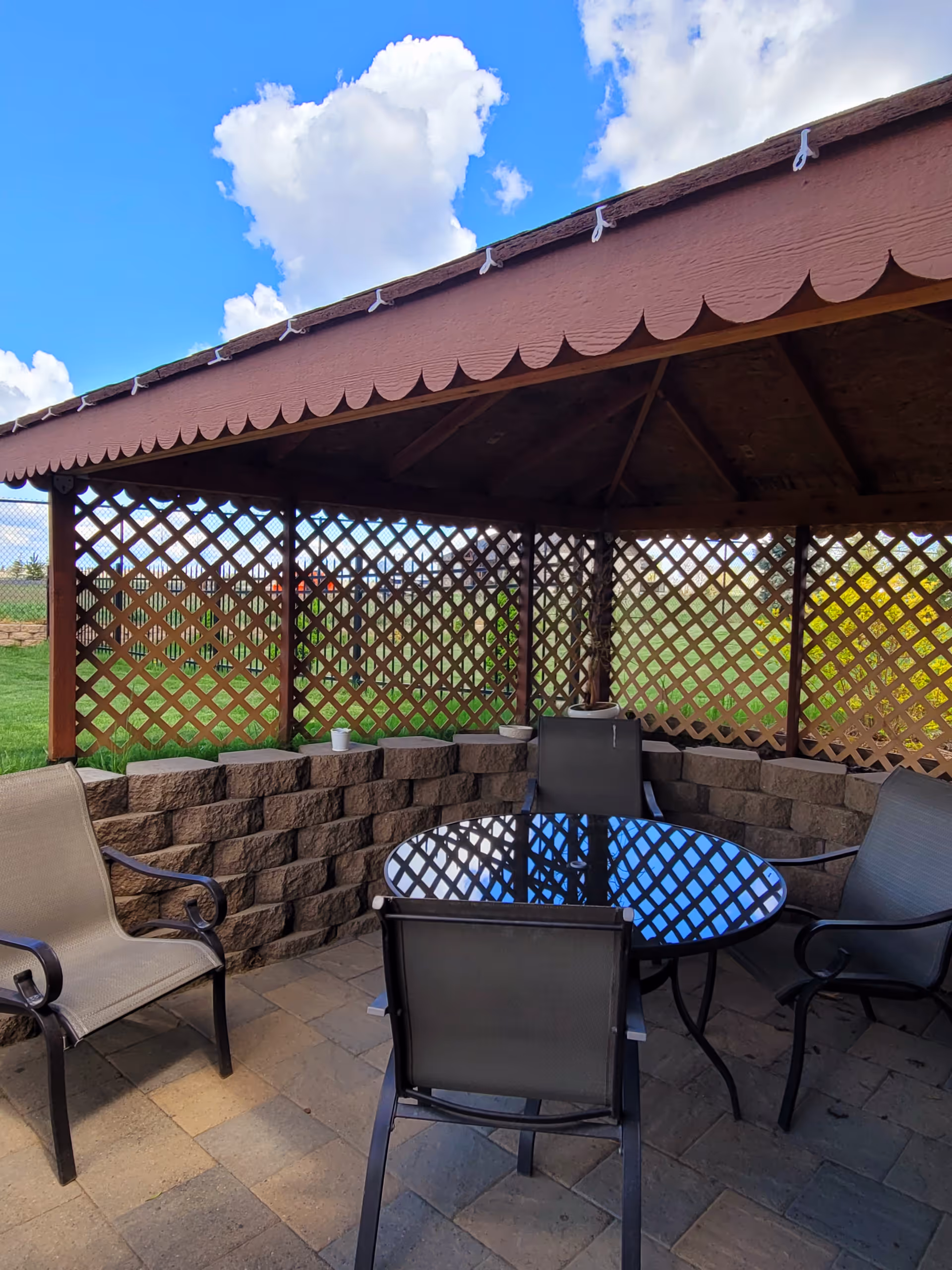 Outdoor seating area under a wooden gazebo with lattice walls, featuring a round glass table and four chairs on a stone-paved floor, with a grassy area and blue sky with clouds visible outside.