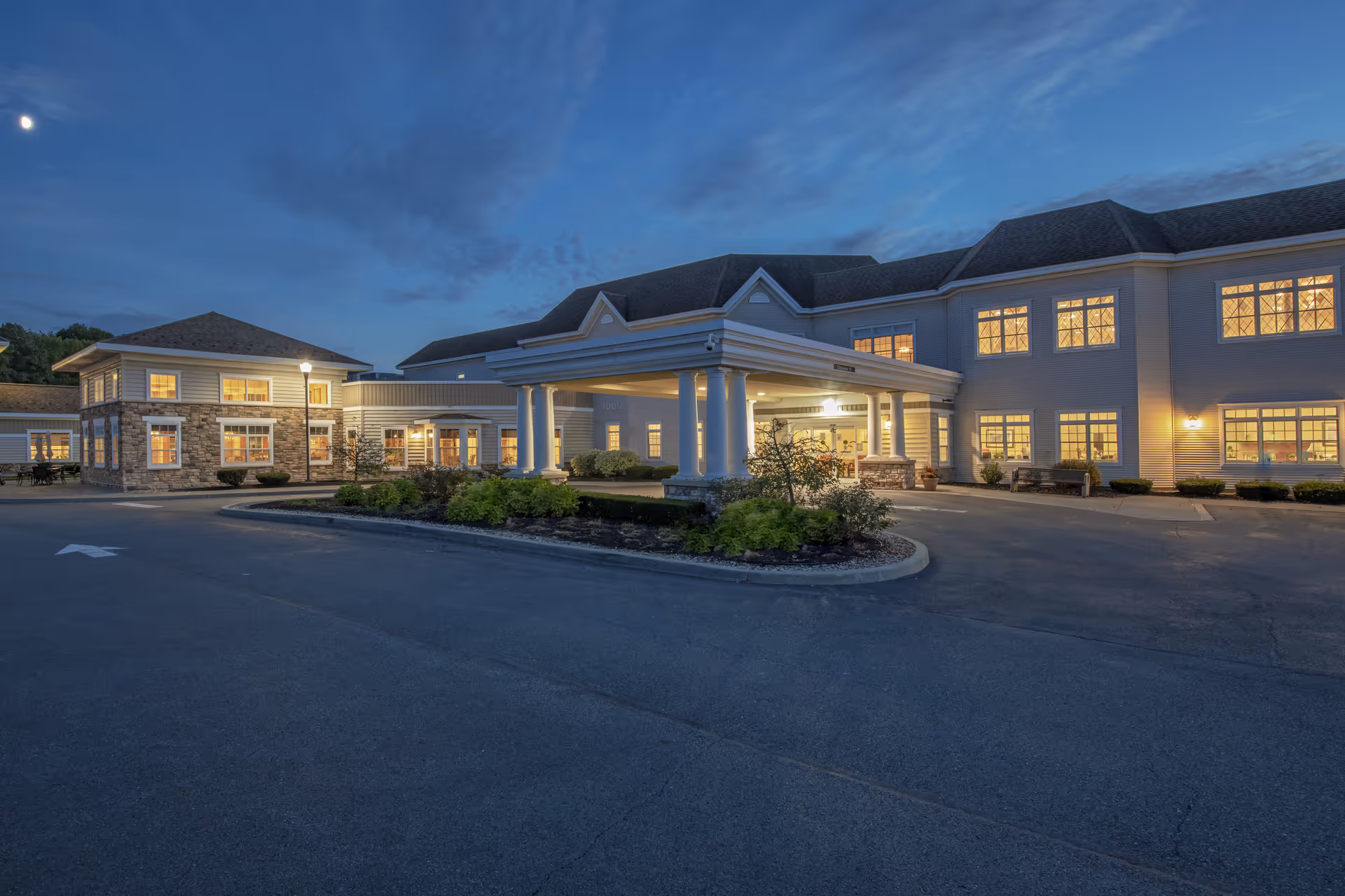 Exterior view of Coburg Village senior living facility at dusk, showing a large building with multiple windows illuminated from inside, a covered entrance with white columns, and a driveway with landscaped greenery in front.