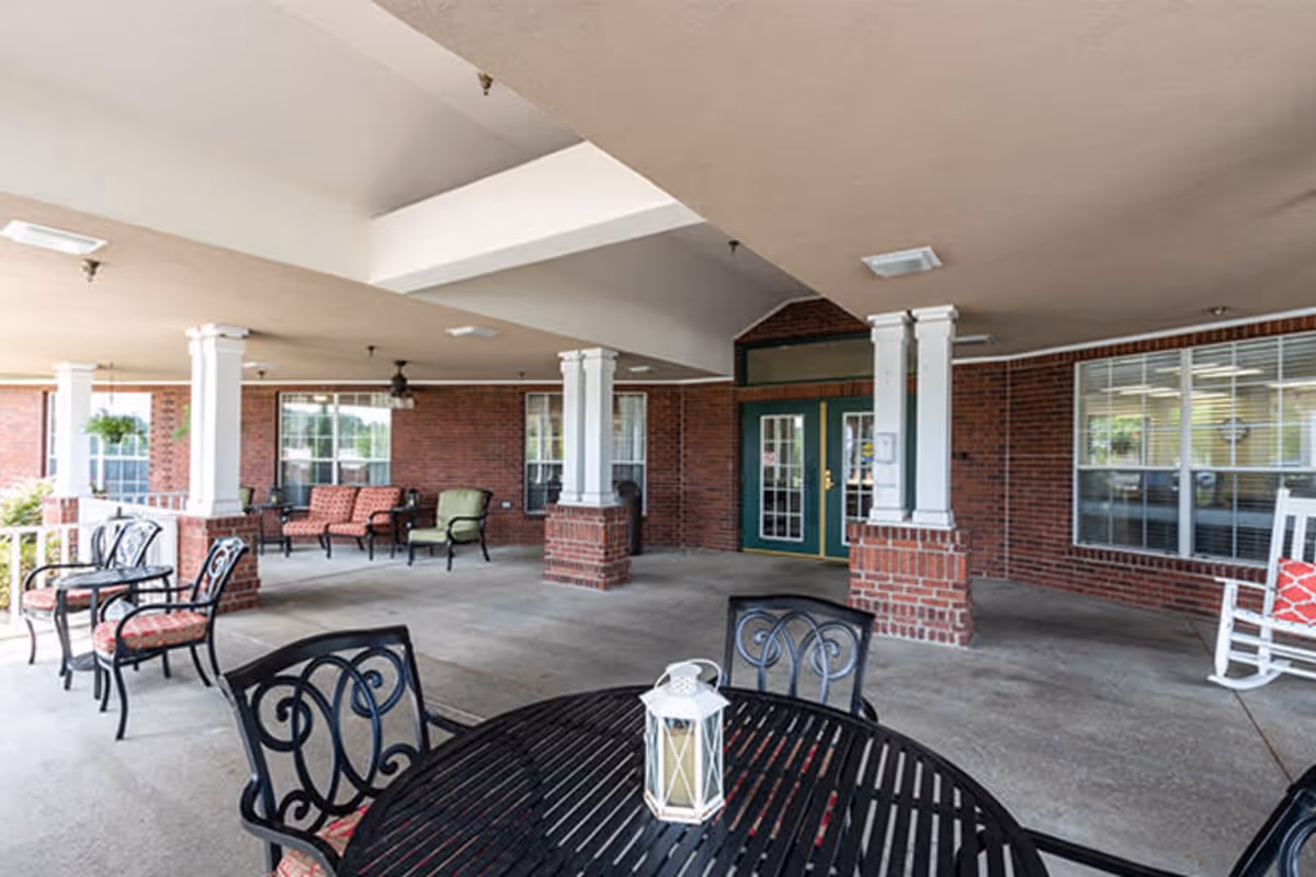Covered outdoor patio area with black metal tables and chairs, cushioned seating, white rocking chair, brick pillars, and green double doors leading inside a building.