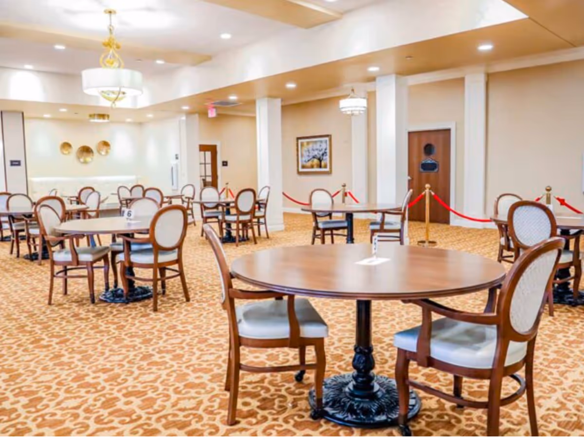 A spacious dining room in a senior living facility with round wooden tables and cushioned chairs arranged neatly on a patterned carpet. The room features warm lighting, decorative wall art, and red velvet ropes sectioning off part of the area.