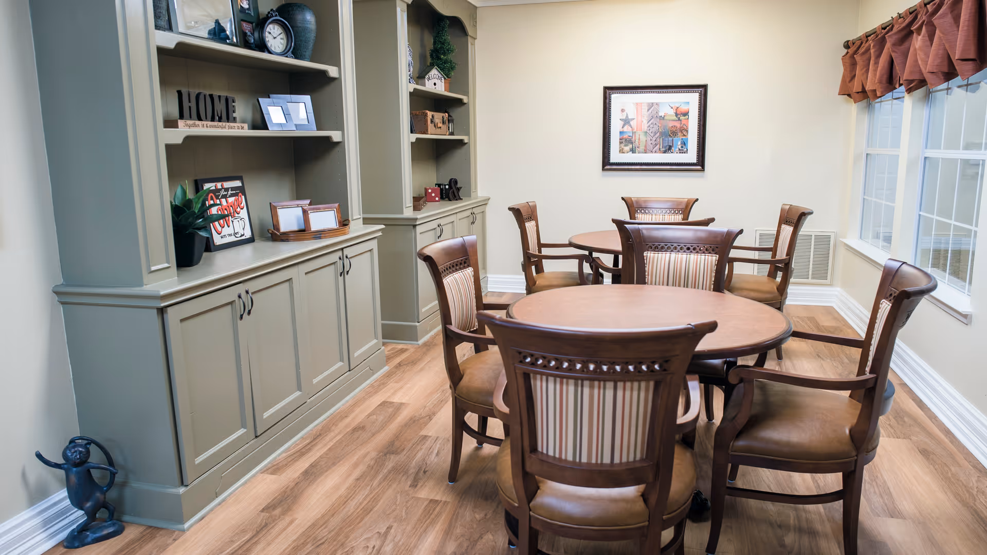 A cozy dining area with two round wooden tables surrounded by wooden chairs with cushioned seats and striped backs. The room features built-in shelves and cabinets painted in a muted green color, decorated with framed pictures, plants, and decorative items. A framed collage artwork hangs on the cream-colored wall, and large windows with brown valances let in natural light. The floor is covered with wood-patterned vinyl or laminate.
