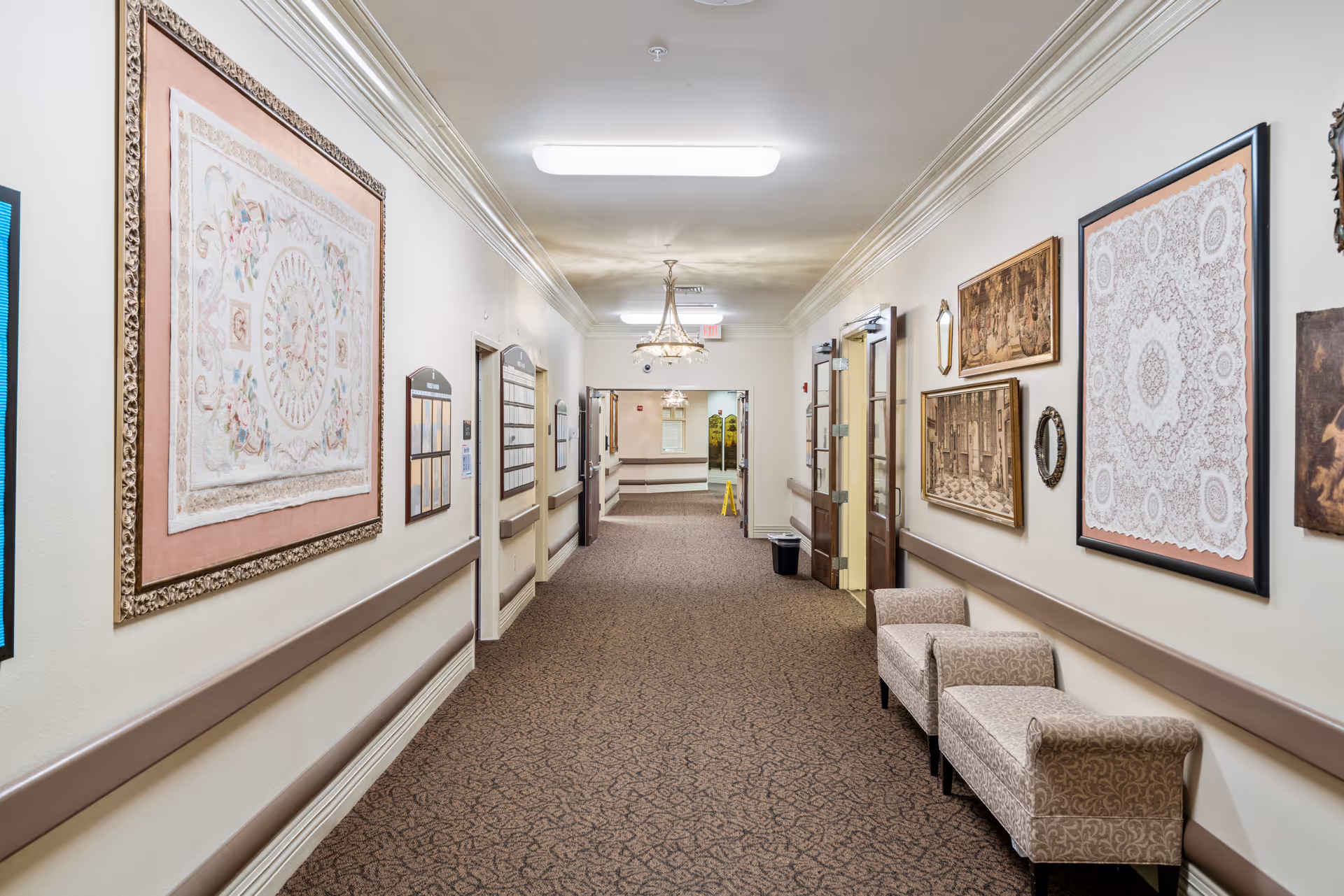A well-lit hallway in a senior living facility with beige walls adorned with framed artwork and decorative pieces. The carpeted floor is brown with a subtle pattern. Two upholstered chairs are placed along the right wall. The hallway has handrails on both sides and doors leading to other rooms. A chandelier and ceiling lights illuminate the space.