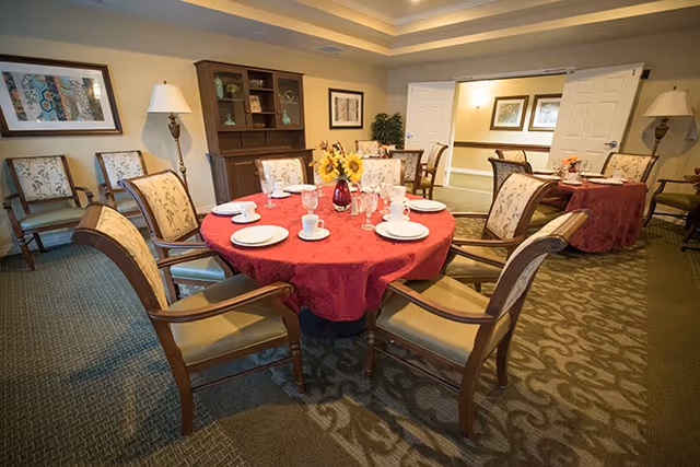 A dining room with round tables covered in red tablecloths, set with white plates, cups, and glassware. The room has wooden chairs with cushioned seats and backs, a wooden cabinet with decorative items, framed artwork on the walls, and soft lighting from table lamps and ceiling lights.
