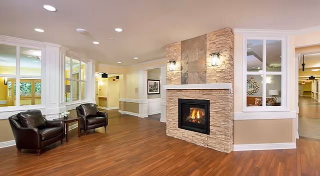 A cozy interior common area featuring a stone fireplace with a fire burning, two dark leather armchairs with a small table between them, wood flooring, and soft lighting. The space has large windows and a hallway leading to other parts of the facility.