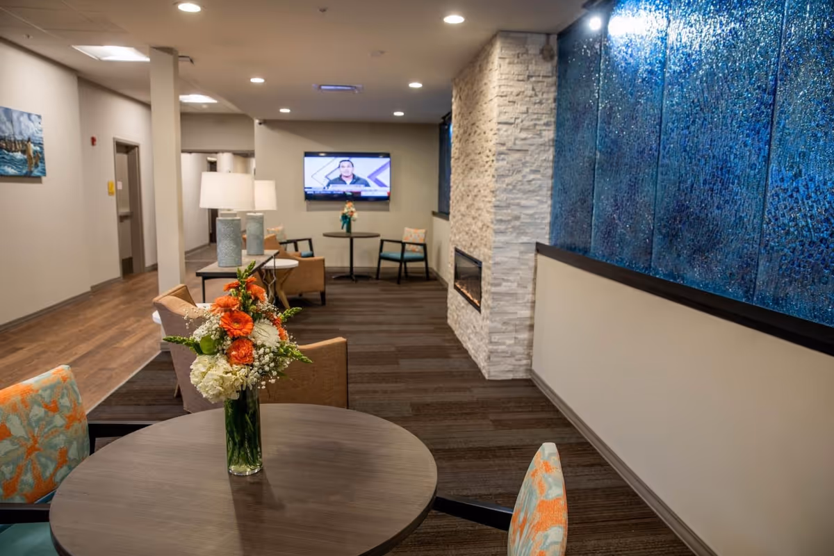 A cozy common area with a round table in the foreground holding a vase of orange and white flowers. There are patterned chairs around the table. The room features a modern fireplace set in a white stone wall and a large blue textured wall art. In the background, there is a wall-mounted TV and additional seating with chairs and a small table. The space has warm lighting and a carpeted floor.