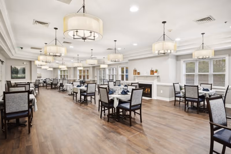 A spacious dining room with multiple tables covered with white tablecloths and set with blue napkins and glassware. The room features wooden flooring, large windows with white blinds, a fireplace with decorative items on the mantel, and several modern pendant lights hanging from the ceiling.