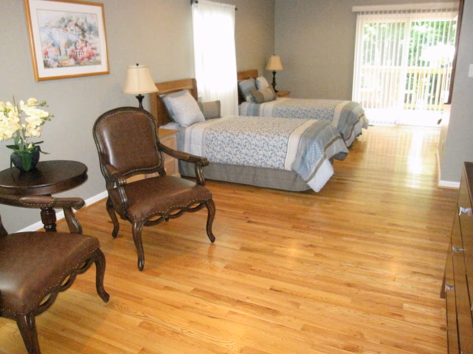 A bedroom with two twin beds covered in blue and white patterned bedding. There are two bedside tables with lamps, a pair of brown leather chairs with a small round table between them, and a framed painting on the wall. The room has wooden flooring and a large window with sheer curtains letting in natural light.