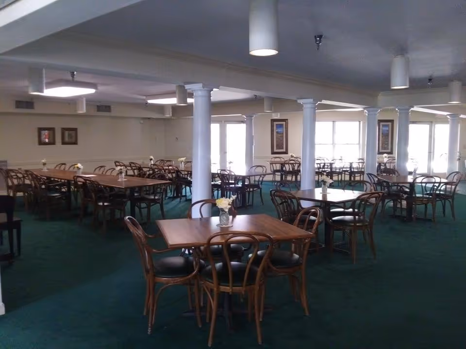 Spacious dining room with multiple wooden tables and chairs, white columns, and green carpet.