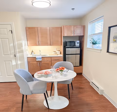 A small kitchen and dining area with wooden cabinets, a microwave, and a mini refrigerator. A round white table with two gray upholstered chairs is set with plates, glasses, and a small flower arrangement. A window with blinds and a framed picture are on the right wall.