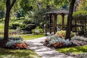 A peaceful outdoor garden area with a paved walkway leading to a wooden gazebo surrounded by trees, shrubs, and colorful flower beds.
