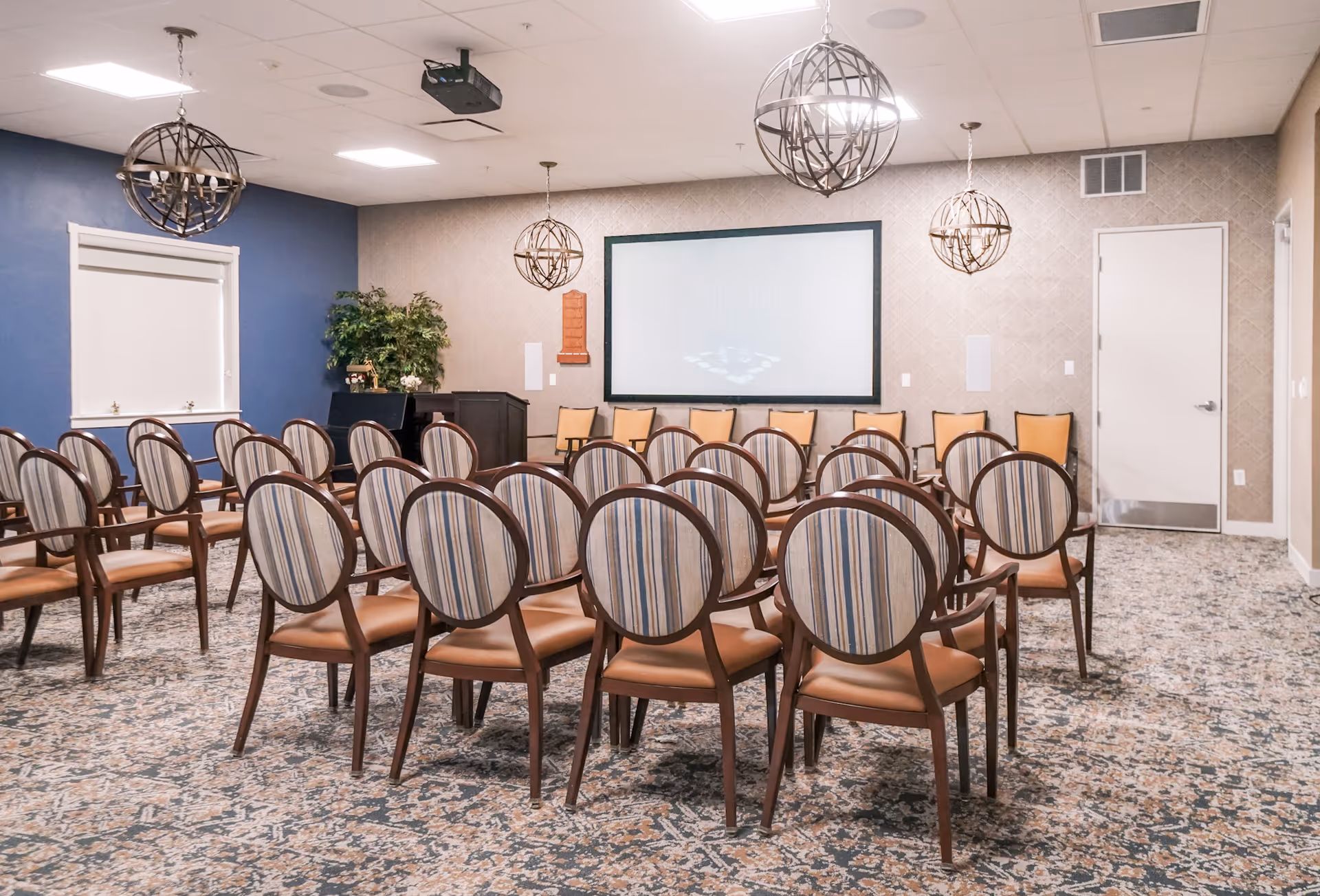 A bright meeting room with rows of striped-upholstered chairs facing a large projection screen and decorative globe pendant lights.