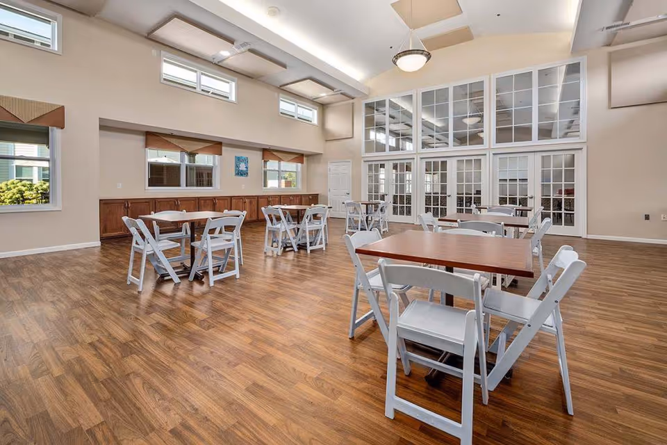 A spacious dining room with wooden floors, several tables with white folding chairs arranged around them, large windows on the walls, and a high ceiling with modern light fixtures.