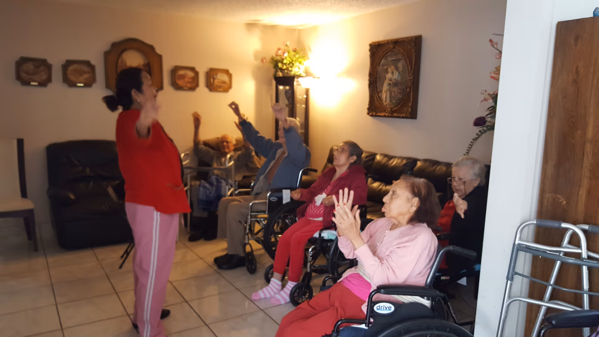 A group of elderly people, some in wheelchairs, participating in a seated exercise or stretching activity led by a woman standing in front of them in a living room setting with tiled floors, leather couches, and wall decorations.