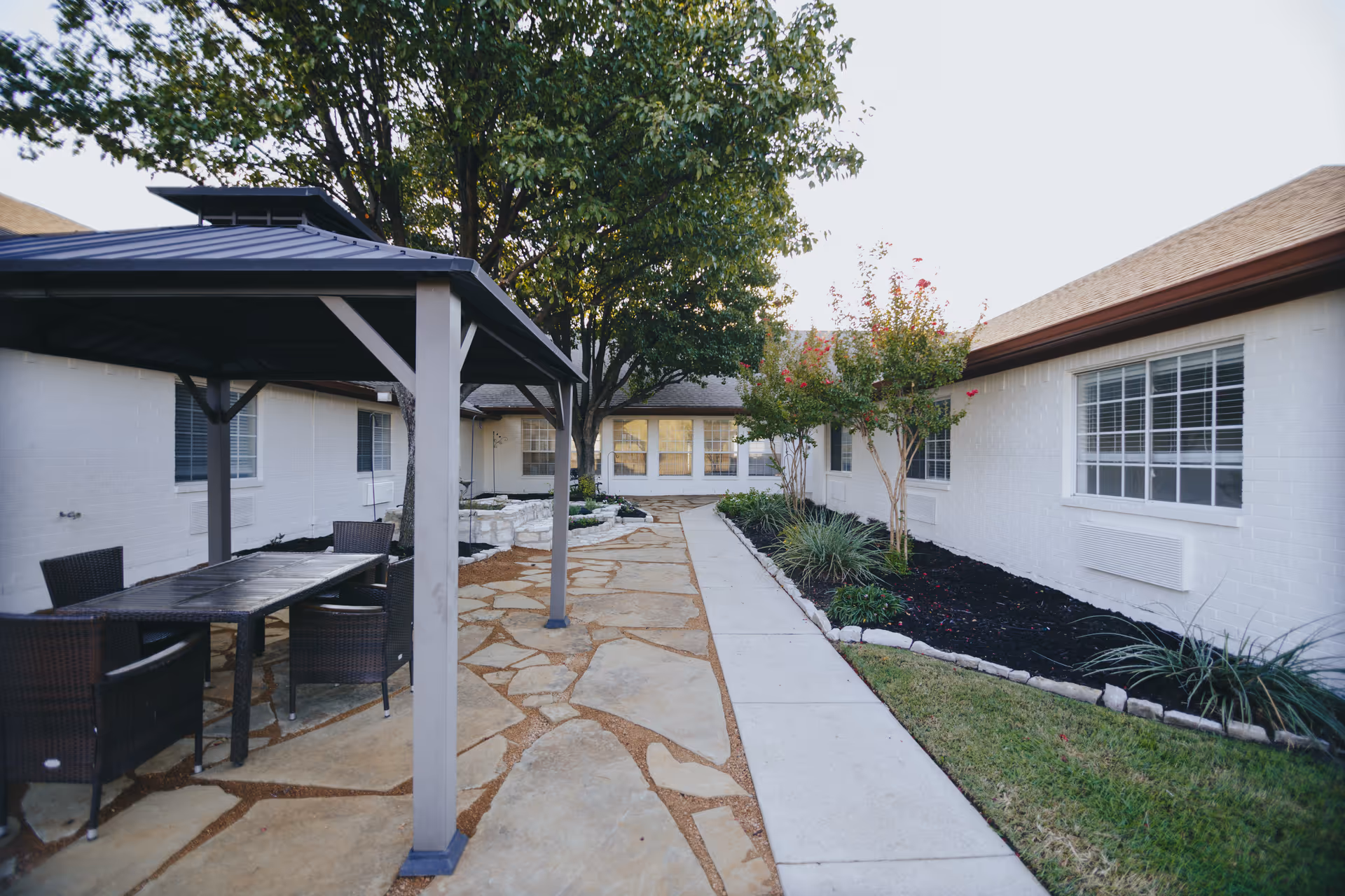 Outdoor courtyard area at Avalon Memory Care featuring a stone-paved walkway, a covered seating area with a table and chairs, trees, and landscaped garden beds alongside white brick buildings with windows.