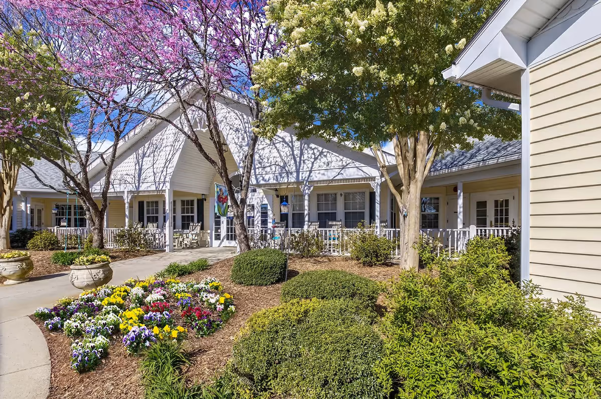 Exterior view of TerraBella Shelby facility showing a well-maintained garden with colorful flowers, shrubs, and trees in bloom. The building has a light yellow siding with white trim and a covered porch with rocking chairs. The sky is clear and blue.