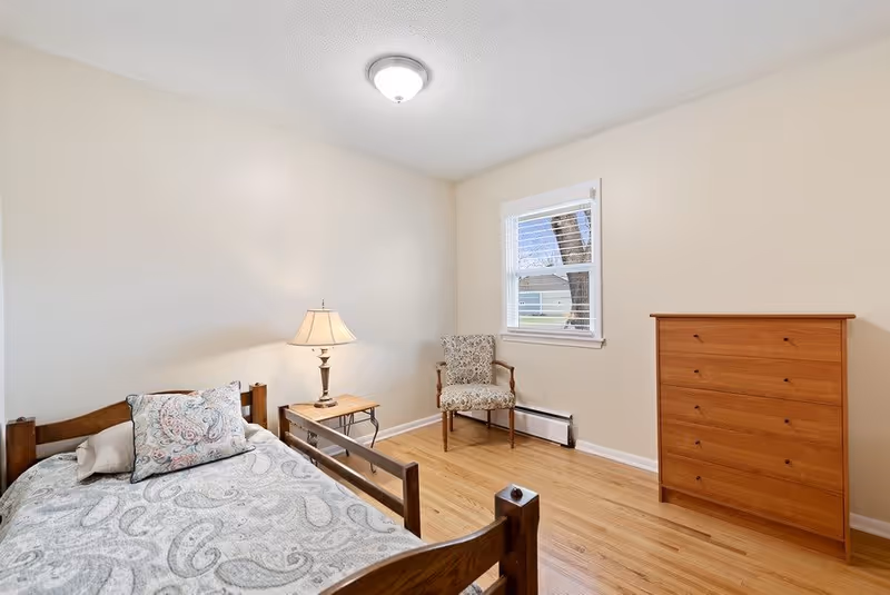 A simple bedroom with a wooden bed frame, a patterned bedspread and pillow, a wooden nightstand with a lamp, a floral upholstered chair near a window with white blinds, and a wooden chest of drawers on a light wooden floor.