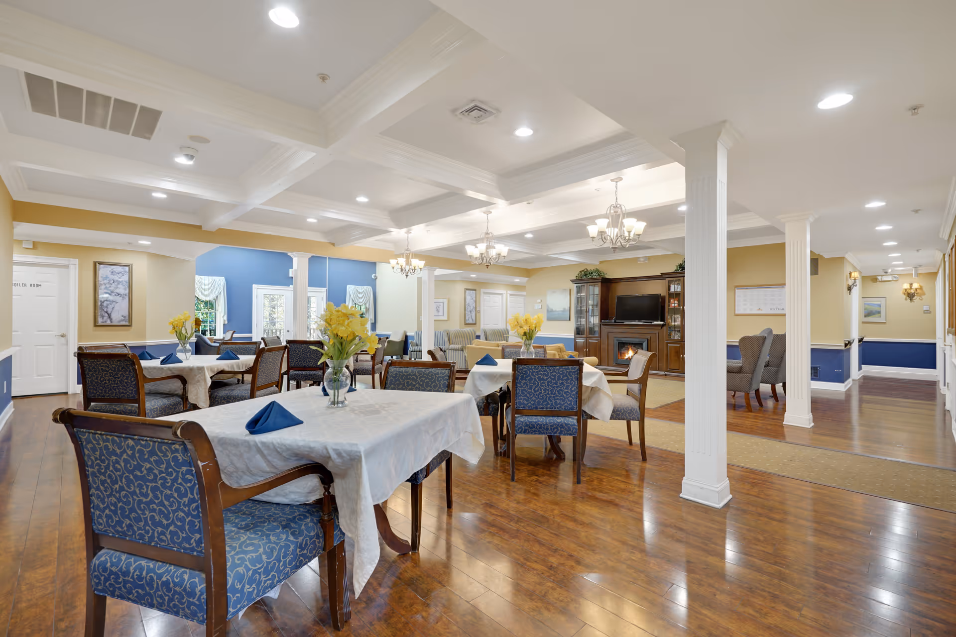A spacious and well-lit dining room with several tables covered in white tablecloths, each adorned with a vase of yellow flowers and blue folded napkins. The room features wooden floors, white coffered ceilings with recessed lighting, and chandeliers. There is a fireplace with a TV above it and comfortable seating areas along the walls. The walls are painted in warm yellow and blue tones, with white columns and trim.