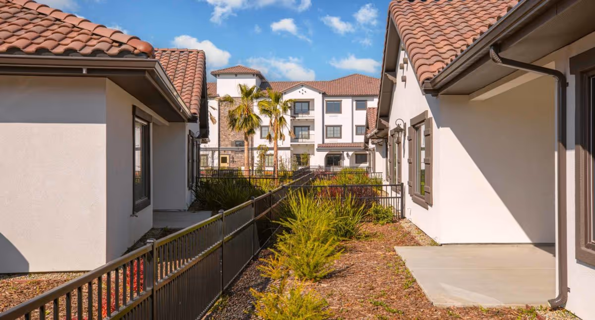 Outdoor view of a senior living facility with white buildings featuring terracotta tiled roofs, a pathway lined with metal railings, landscaped plants, and palm trees under a blue sky with scattered clouds.