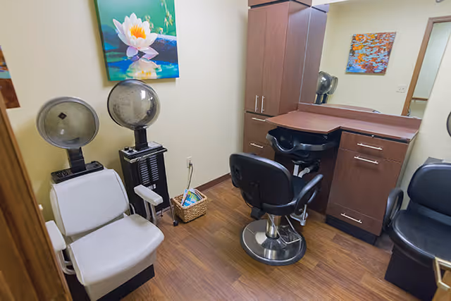 Interior view of a small salon room with two vintage hair dryers, a salon chair in front of a counter with a mirror, wooden cabinets, and a basket with magazines on the floor. A colorful painting of a water lily is hung on the wall.